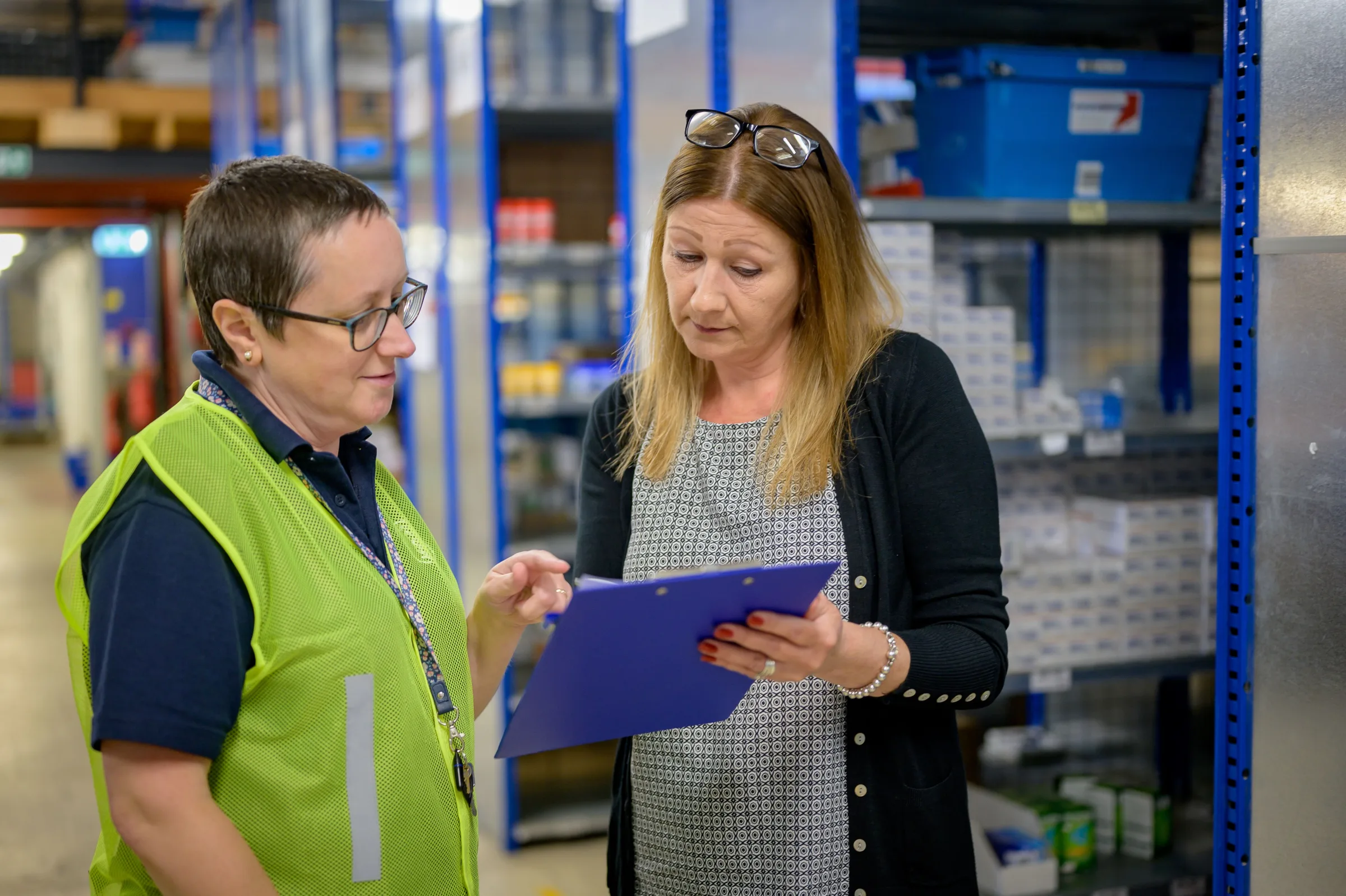 Two workplace colleagues reviewing a clipboard beside warehouse shelving.